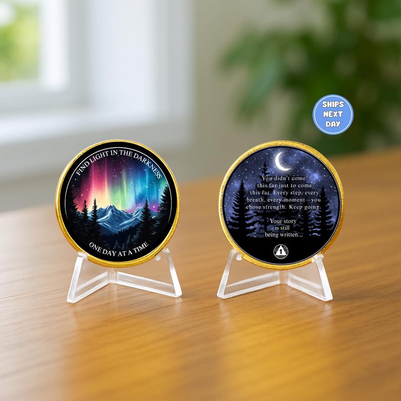 May include: Two gold-rimmed coins on clear stands. One coin features a colorful aurora borealis scene with text: "FIND LIGHT IN THE DARKNESS, ONE DAY AT A TIME." The other coin has a night sky scene with text: "You didn't come this far... Your story is still being written..."
