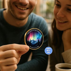 May include: A round, gold-rimmed coin featuring a colorful aurora borealis scene above a mountain range and forest. The coin has the text "FIND LIGHT IN THE DARKNESS" and "ONE DAY AT A TIME".