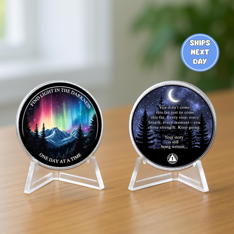 May include: Two round, silver-colored coins on clear stands. One coin features a colorful aurora borealis scene with text: "FIND LIGHT IN THE DARKNESS" and "ONE DAY AT A TIME." The other coin has a night sky scene with a crescent moon and text: "You didn't come this far..."
