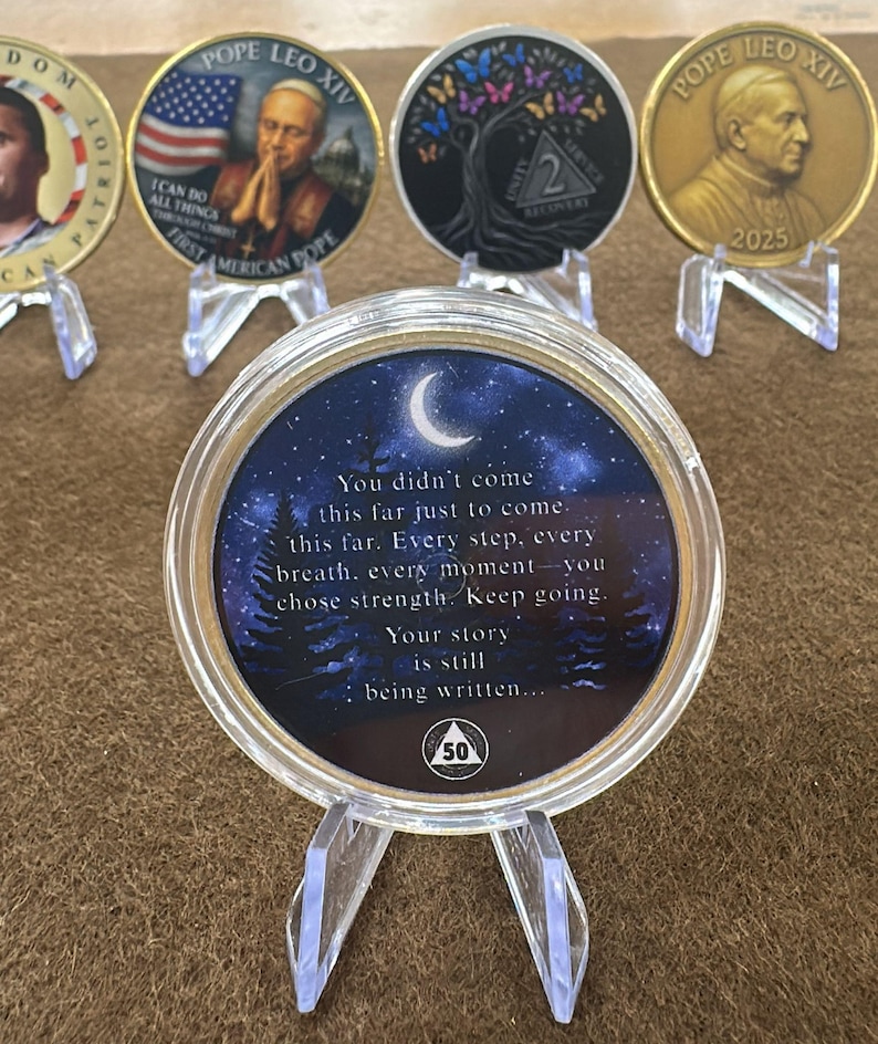 May include: A collection of commemorative coins displayed on clear stands. The coins feature various designs, including portraits, flags, and inspirational quotes. One coin has the text "You didn't come this far just to come..." and a crescent moon.