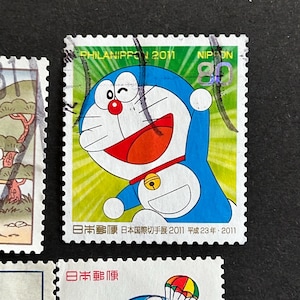 May include: A postage stamp featuring the cartoon character Doraemon, with a blue body, white face, and red mouth. The stamp has a green background with the text "PHILANIPPON 2011" and "NIPON 80". Japanese text is also present.