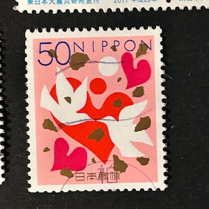 May include: A Japanese postage stamp with a pink background features a design of two white doves, red hearts, and a red abstract shape. The stamp also includes the text "50 NIPPON" in blue and Japanese characters.