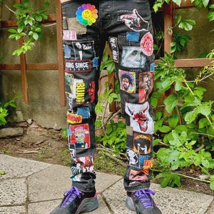 Può includere: Jeans in denim nero caratterizzati da una varietà di toppe colorate, tra cui un fiore arcobaleno con una faccina sorridente, un ritratto di Benjamin Franklin, un teschio di diavolo rosso e varie stampe grafiche, indossati con scarpe da ginnastica nere e viola.
