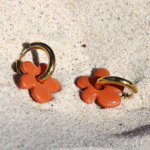 Può includere: Un paio di orecchini a forma di fiore arancione con cerchi dorati. Gli orecchini sono appoggiati su una superficie sabbiosa. Un accessorio alla moda dal design accattivante.