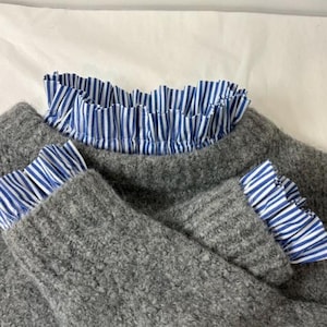 Può includere: Un maglione grigio con volant a righe blu e bianche sul colletto e sui polsini. Il maglione ha un aspetto a maglia strutturata e i volant aggiungono un dettaglio a contrasto.