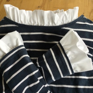 Puede incluir: Una camisa de manga larga a rayas azul marino y blancas con ribete blanco con volantes alrededor del cuello y los puños.