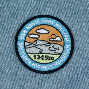 May include: A circular embroidered patch featuring a mountain landscape on light blue denim fabric. The patch displays the text BEN NEVIS FORT WILLIAM BRITAIN'S HIGHEST MOUNTAIN and 1345m, with a graphic of a mountain, clouds, and a sun.