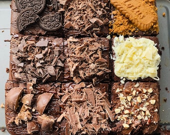 May include: A tray of nine chocolate brownies, each topped with a different ingredient. The brownies are arranged in a 3x3 grid. The toppings include chocolate chips, chocolate shavings, white chocolate shavings, chopped nuts, and crumbled cookies.
