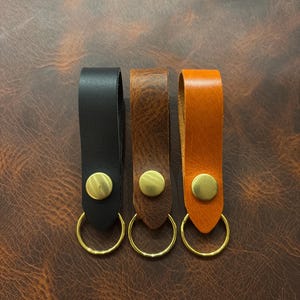 Op de afbeelding: Drie leren sleutelhangers in zwart, bruin en oranje. Elke sleutelhanger heeft een messingkleurige drukknop en een goudkleurige sleutelring. De sleutelhangers worden gepresenteerd op een bruin leren oppervlak.