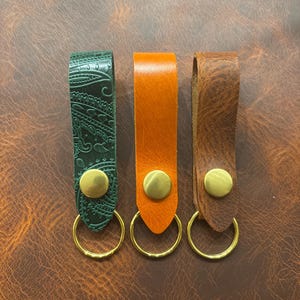 Op de afbeelding: Drie leren sleutelhangers in groen, oranje en bruin. De groene sleutelhanger heeft een paisley patroon. Elke sleutelhanger heeft een goudkleurige metalen ring en een bijpassende klinknagel. De sleutelhangers worden gepresenteerd op een bruin leren oppervlak.