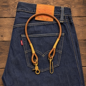 Könnte beinhalten: Eine braune Geldbörsenkette aus Leder mit gelben Garnakzenten und Messingbeschlägen, einschließlich eines drehbaren Karabinerhakens und eines Hakens, auf einer dunkelblauen Jeans mit einem hellbraunen Lederaufnäher von Levi Strauss & Co.