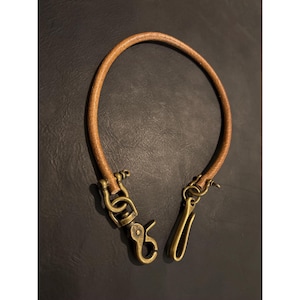 Op de afbeelding: Een bruine leren portemonnee ketting met messingkleurige hardware. De ketting is cirkelvormig en heeft aan één uiteinde een sluiting en aan het andere uiteinde een haak. Het leer heeft een lichtbruine kleur.