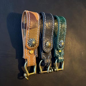 May include: Three leather keychains in shades of brown, black, and green. Each keychain features a decorative metal stud and a brass-colored clasp. The brown keychain has a camouflage pattern, while the black and green ones have embossed designs.