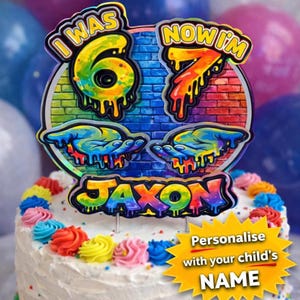May include: A circular cake topper featuring a brick wall background with a rainbow gradient. The text reads I WAS 6 NOW I'M 7 and JAXON. The design includes two hands and numbers with a dripping paint effect in vibrant yellow, orange, and blue.