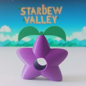 May include: A purple, star-shaped object with a central hole and two green leaf-like elements on top. The object is a stylized representation of a crop from the video game Stardew Valley. The background features the game's logo and a landscape.
