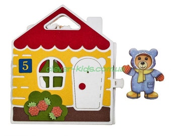 Könnte beinhalten: Ein gelbes Filzhaus mit einem roten Dach, einer weißen Tür mit einem roten Kreis und einem Fenster. Das Haus hat die Nummer "5" an der Seite. Ein blauer Filzbär mit einem gelben Schal und orangenen Fäustlingen steht neben dem Haus.