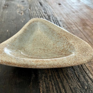 May include: A triangular ceramic dish with a speckled glaze in shades of beige and brown. The dish has a slightly raised rim and a smooth, glossy finish.