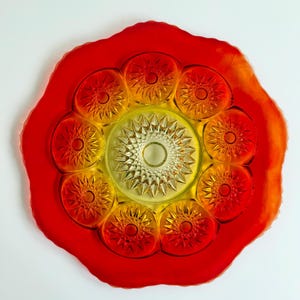 May include: A round glass serving plate featuring a vibrant red outer border and a yellow center. The plate is decorated with a series of pressed glass starburst patterns arranged in a circular formation around a central starburst motif.