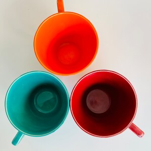 Puede incluir: Tres tazas de cer&aacute;mica en diferentes colores: naranja, verde azulado y granate. Las tazas est&aacute;n vac&iacute;as y tienen un fondo blanco.