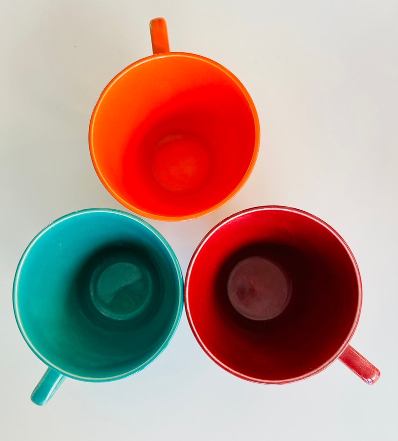 Puede incluir: Tres tazas de cer&aacute;mica en diferentes colores: naranja, verde azulado y granate. Las tazas est&aacute;n vac&iacute;as y tienen un fondo blanco.