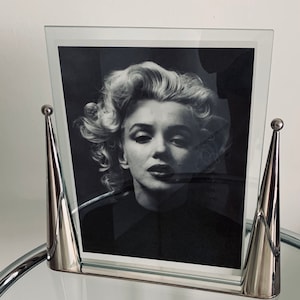May include: A silver glass picture frame with a black and white photo of Marilyn Monroe. The frame has two silver cone-shaped bases.