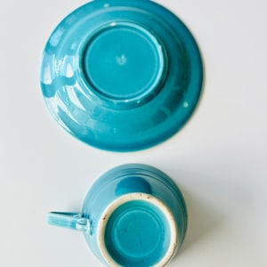 Puede incluir: Un juego de taza y platillo de cer&aacute;mica azul turquesa. La taza tiene un asa y el platillo tiene un borde ligeramente elevado.