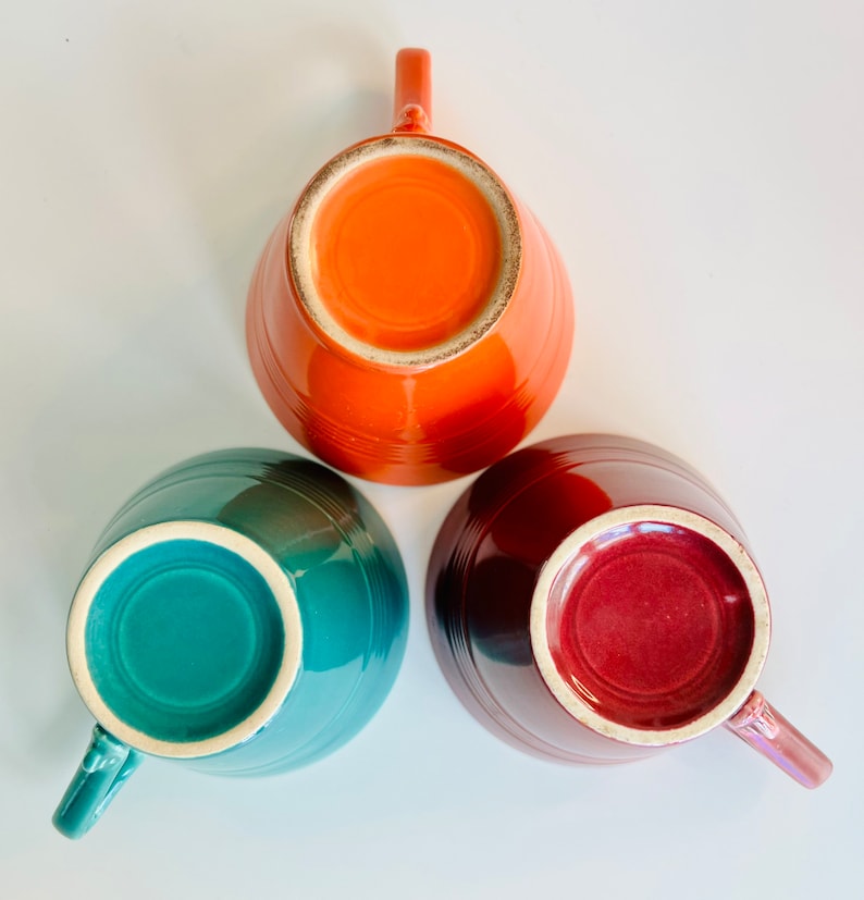 Puede incluir: Tres tazas de cer&aacute;mica en diferentes colores: verde azulado, naranja y granate. Las tazas est&aacute;n boca abajo, mostrando el interior de las tazas.