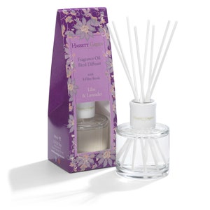 May include: A clear glass fragrance oil reed diffuser with white reeds and a white top, next to a purple box. The box has floral designs and text that reads "Lilac & Lavender" and "Fragrance Oil Reed Diffuser".