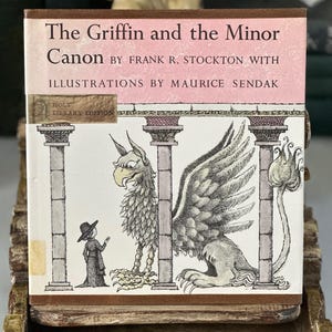 May include: A vintage book titled "The Griffin and the Minor Canon" by Frank R. Stockton, with illustrations by Maurice Sendak. The cover features a detailed illustration of a griffin and a person. The book is displayed on a decorative wooden stand.