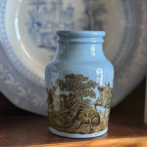 May include: A small, light blue ceramic vase with a detailed, sepia-toned relief depicting a hunting scene. The vase has a slightly flared rim and a cylindrical body. A blue and white plate is in the background.