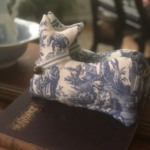 May include: A blue and white toile patterned fabric stuffed animal in the shape of a dog lying on top of a stack of books. The books have gold lettering on the spines.