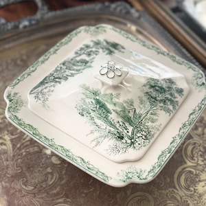 May include: A white and green ceramic serving dish with a lid. The dish features a green landscape design and a decorative green border. The lid has a small, flower-shaped handle. The dish is square with rounded corners.