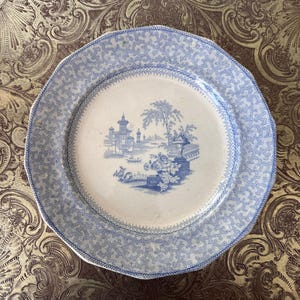 May include: A round, antique plate with a blue and white design. The plate features a detailed landscape scene in the center, surrounded by a patterned border. The plate is likely made of ceramic or porcelain and is a vintage item.