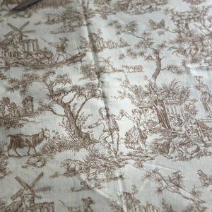 May include: Cream-colored fabric with a brown toile pattern depicting pastoral scenes, windmills, and figures. Gold scissors and a checkered fabric are visible at the top. This fabric is suitable for various sewing projects.