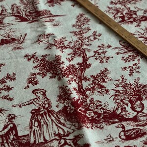 May include: Cream fabric featuring a red toile design with pastoral scenes, figures, trees, and bridges. A wooden ruler, marked in inches, lies diagonally across the fabric. The fabric is suitable for various craft projects.
