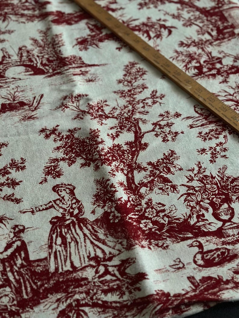 May include: Cream fabric featuring a red toile design with pastoral scenes, figures, trees, and bridges. A wooden ruler, marked in inches, lies diagonally across the fabric. The fabric is suitable for various craft projects.