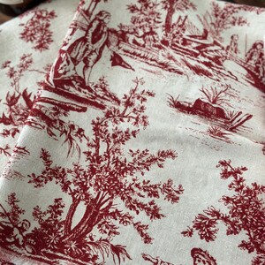 May include: Cream-coloured fabric featuring a red toile de Jouy design with figures in a rural setting. The fabric has a fringed edge, and the pattern includes trees, people, and buildings.
