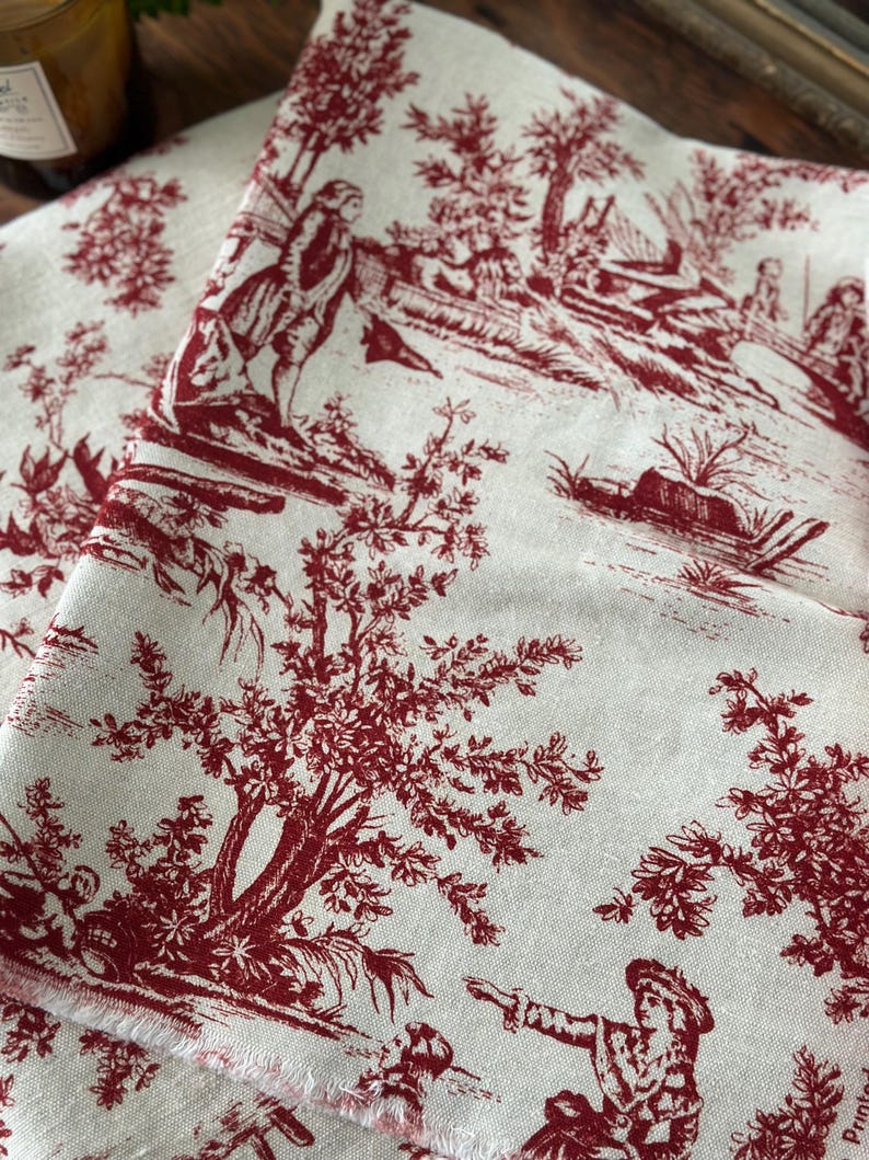 May include: Cream-coloured fabric featuring a red toile de Jouy design with figures in a rural setting. The fabric has a fringed edge, and the pattern includes trees, people, and buildings.