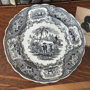 May include: A decorative plate with a black and white transferware design. The plate features a central scene of people near a body of water with palm trees and a ship, surrounded by floral and landscape motifs. The plate has a scalloped edge.