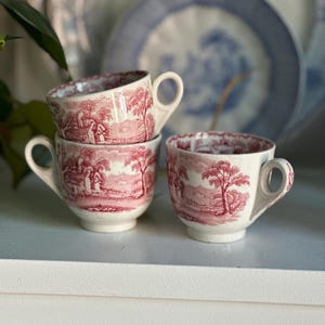 May include: Four white teacups with a red pastoral scene design. The cups are stacked and arranged on a white surface, with a blurred blue and white plate in the background. The teacups have a classic, vintage aesthetic.