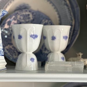 May include: Two white porcelain egg cups with blue floral designs. The egg cups are on a white shelf, with a blue and white patterned plate in the background. The egg cups are part of a larger collection of blue and white porcelain.