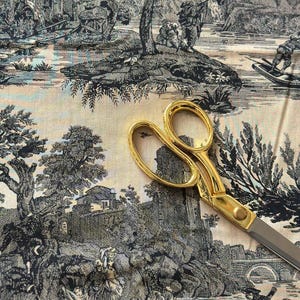 May include: Gold-handled scissors rest on a cream-colored fabric with a black and white toile pattern depicting a landscape with figures, trees, and buildings. The scissors have silver blades.