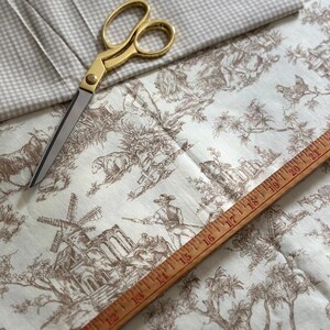 May include: A collection of fabric and sewing tools. The image features a pair of gold scissors, a wooden ruler, and two types of fabric. One fabric has a brown toile pattern, and the other is a light gray gingham check.