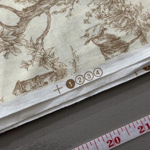 May include: Cream-colored fabric with a brown pastoral toile pattern featuring trees, buildings, and animals. The fabric is folded, with a measuring tape showing inches. Numbers 1, 2, 3, and 4 are printed on the fabric.