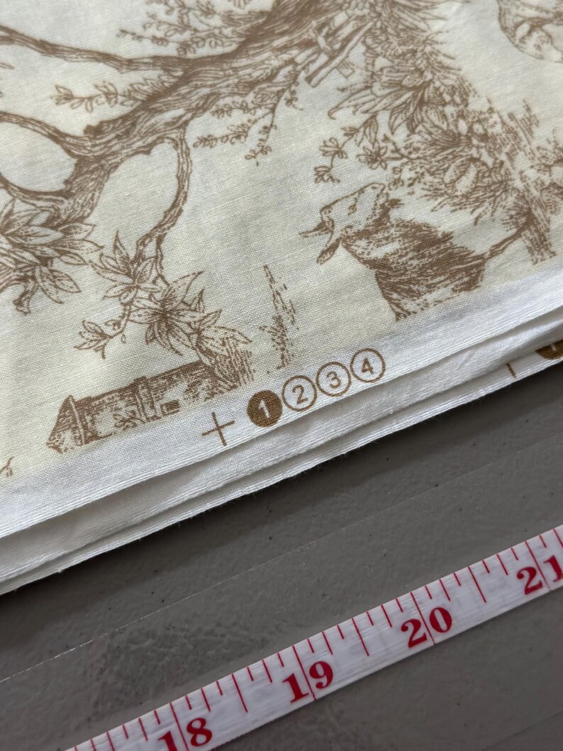 May include: Cream-colored fabric with a brown pastoral toile pattern featuring trees, buildings, and animals. The fabric is folded, with a measuring tape showing inches. Numbers 1, 2, 3, and 4 are printed on the fabric.