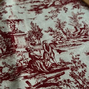 May include: A close-up of fabric featuring a red and white toile de Jouy design. The pattern shows detailed scenes of people, trees, and buildings. The fabric has a soft texture and a classic aesthetic.