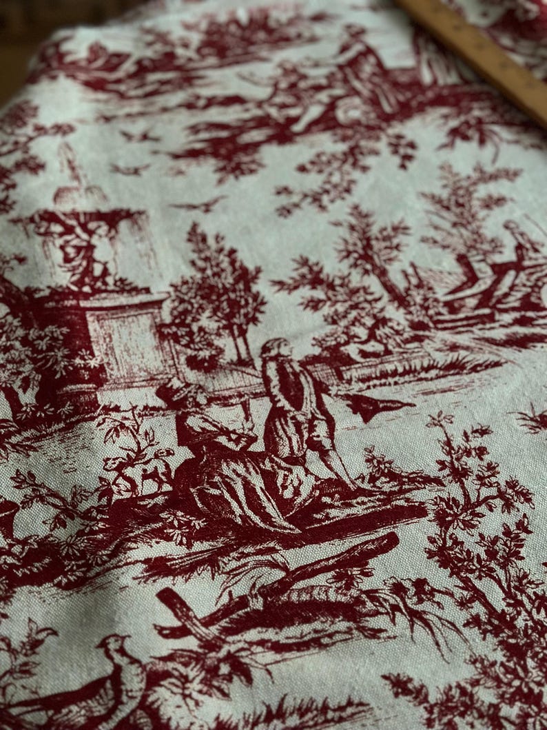 May include: A close-up of fabric featuring a red and white toile de Jouy design. The pattern shows detailed scenes of people, trees, and buildings. The fabric has a soft texture and a classic aesthetic.