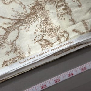 May include: Cream-colored fabric with a brown toile pattern featuring figures, trees, and buildings. The fabric has the text "Classic Cottons &copy; 2006 Visit www.classiccottons.com". A measuring tape with inch markings is visible.