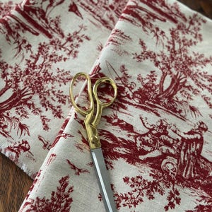 May include: Cream fabric with a red toile de Jouy design depicting rural scenes. Gold-handled scissors with silver blades are placed on the fabric. The fabric has a raw, unfinished edge.