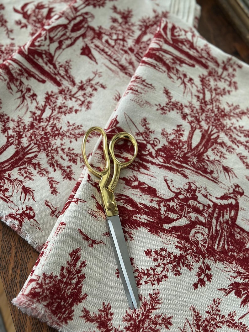 May include: Cream fabric with a red toile de Jouy design depicting rural scenes. Gold-handled scissors with silver blades are placed on the fabric. The fabric has a raw, unfinished edge.